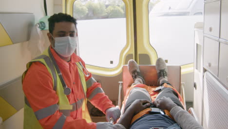 Portrait-Of-A-Male-Paramedic-With-Face-Mask-Looking-At-The-Camera-While-Riding-In-An-Ambulance-And-Taking-Care-Of-An-Injured-American-Patient