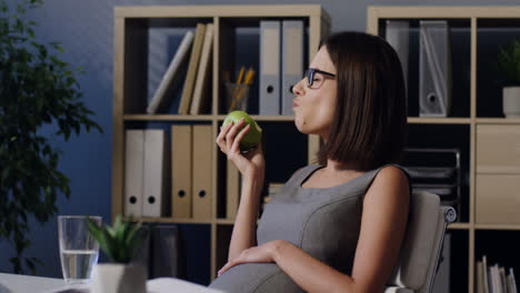 Mujer-Embarazada-Con-Gafas-Sentada-En-La-Oficina-Comiendo-Fruta-Y-Bebiendo-Agua