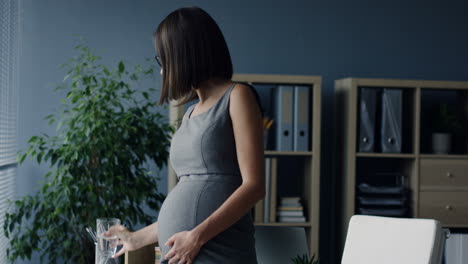 Schwangere-Geschäftsfrau,-Die-Kopfschmerzen-Hat-Und-Wasser-Trinkt,-Während-Sie-Im-Büro-Steht