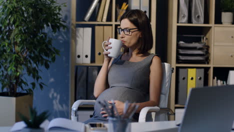 Pregnant-Businesswoman-In-Glasses-Caressing-Her-Belly-And-Drinking-Tea-Sitting-On-A-Chair-In-The-Office