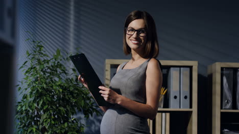 Schwangere-Geschäftsfrau-Mit-Brille-Liest-Dokumente-Im-Büro-Und-Lächelt-Dann-In-Die-Kamera