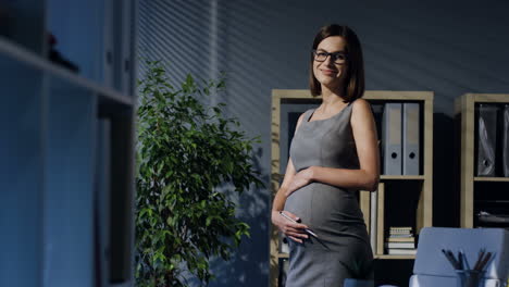 Schwangere-Geschäftsfrau-Mit-Brille-Streichelt-Ihren-Bauch-Im-Büro-Und-Lächelt-Dann-In-Die-Kamera