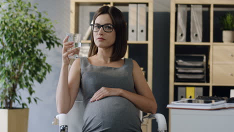 Mujer-De-Negocios-Embarazada-Con-Gafas-Descansando-En-La-Silla,-Bebiendo-Agua-Y-Sonriendo-A-La-Cámara-En-La-Oficina