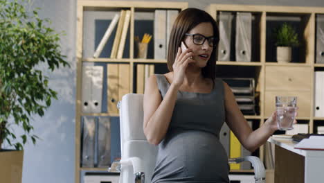 Schwangere-Geschäftsfrau-Mit-Brille,-Die-Im-Büro-Sitzt-Und-Telefoniert,-Während-Sie-Wasser-Trinkt