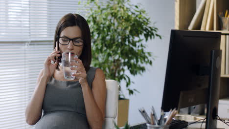 Schwangere-Geschäftsfrau-Mit-Brille,-Die-Im-Büro-Sitzt-Und-Telefoniert,-Während-Sie-Wasser-Trinkt-1