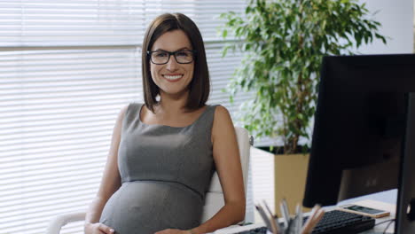 Schwangere-Geschäftsfrau-Mit-Brille-Sitzt-Am-Tisch-Im-Büro-Und-Lächelt-In-Die-Kamera,-Während-Sie-Ihren-Bauch-Streichelt