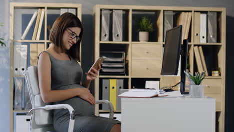 Schwangere-Geschäftsfrau-Mit-Brille,-Die-Am-Schreibtisch-Im-Büro-Sitzt,-Während-Sie-Das-Smartphone-Benutzt-Und-Ihren-Bauch-Streichelt