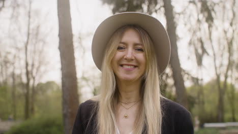 Portrait-Of-A-Happy-Blonde-Woman-With-Hat-Standing-In-The-Park-And-Looking-At-The-Camera
