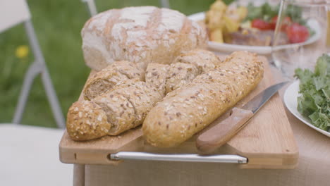 Close-Up-Shot-Of-Fresh-Bread-On-A-Wooden-Board-On-A-Dining-Table-For-An-Outdoor-Party-In-The-Park