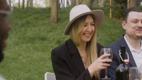 Blond-Woman-Holding-A-Red-Wine-Glass-And-Laughing-While-Sitting-At-Table-With-Friends-During-An-Outdoor-Party-In-The-Park