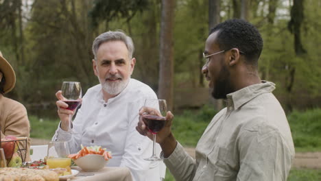 Two-Middle-Aged-Friends-Drinking-Red-Wine-While-Sitting-At-Table-During-An-Outdoor-Party-In-The-Park