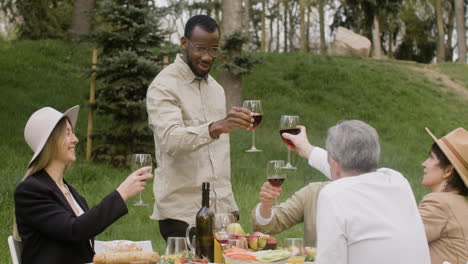 Man-Standing-In-Front-Of-A-Table-Toasting-With-His-Friends-At-An-Outdoor-Party-In-The-Park