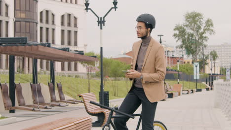 Junger-Amerikaner-In-Formeller-Kleidung-Und-Helm-Mit-Handy,-Während-Er-Auf-Dem-Fahrrad-In-Der-Stadt-Sitzt