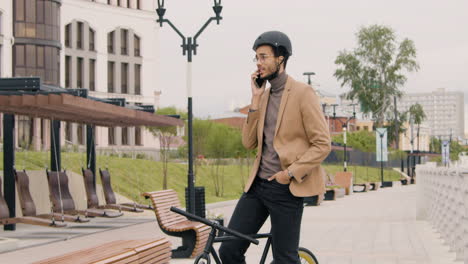 Young-American-Man-In-Formal-Clothes-And-Helmet-Having-A-Call-On-Mobile-Phone-While-Sitting-On-Bike-In-The-City