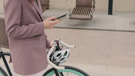 Nahaufnahme-Einer-Nicht-Erkennbaren-Frau-In-Formeller-Kleidung,-Die-Mit-Einem-Fahrrad-Geht-Und-Ein-Handy-In-Der-Stadt-Benutzt