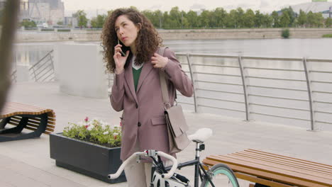 Lockige-Frau-In-Formeller-Kleidung-Und-Fahrrad,-Die-Auf-Der-Stadtbrücke-Mit-Dem-Handy-Telefonieren