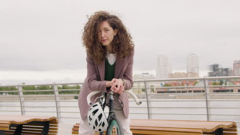 Beautiful-Curly-Woman-In-Formal-Clothes-Looking-And-Smiling-At-The-Camera-While-Sitting-On-A-Bicyle-On-The-City-Bridge