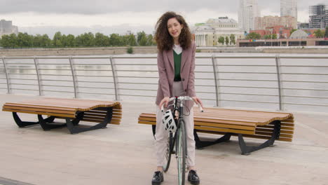 Schöne-Lockige-Frau-In-Formeller-Kleidung,-Die-Auf-Einem-Fahrrad-Auf-Der-Stadtbrücke-1-Sitzt-Und-In-Die-Kamera-Lächelt