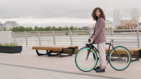 Beautiful-Curly-Woman-In-Formal-Clothes-Looking-And-Smiling-At-The-Camera-While-Sitting-On-A-Bicyle-On-The-City-Bridge-2