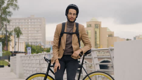 Handsome-American-Man-In-Formal-Clothes-With-Helmet-And-Backpack-Looking-And-Smiling-At-The-Camera-While-Leaning-On-A-Bicyle-On-The-City-Bridge-1