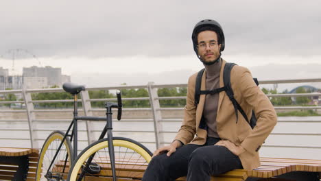 Handsome-American-Man-In-Formal-Clothes-With-Helmet-And-Backpack-Looking-At-The-Camera-While-Sitting-Next-To-His-Bike-On-A-Bench-On-The-City-Bridge
