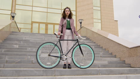 Serious-Curly-Woman-In-Formal-Clothes-Looking-At-The-Camera-While-Standing-On-The-Stairs-With-Her-Bike