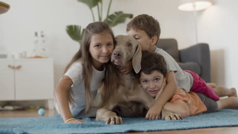 Happy-Kids-Lying-On-Floor-And-Hugging-A-Lovely-Dog-At-Home-And-Looking-At-The-Camera
