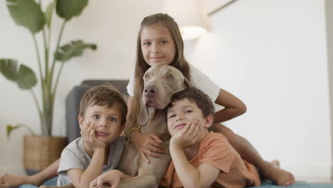 Cheerful-Kids-Lying-With-Dog-On-Floor-And-Looking-At-Camera