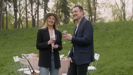 Middle-Aged-Man-And-Woman-Laughing-And-Looking-At-Camera-While-Holding-Wine-Glasses-In-The-Park