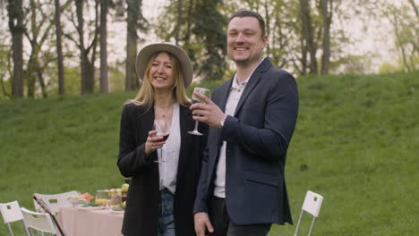 Middle-Aged-Man-And-Woman-Laughing-And-Looking-At-Camera-While-Toasting-With-Wine-Glasses-In-The-Park