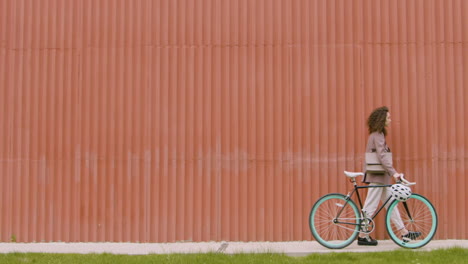 Mujer-Joven-Con-Ropa-Formal-Caminando-Con-Bicicleta-Frente-A-Un-Edificio-De-Metal-Prefabricado