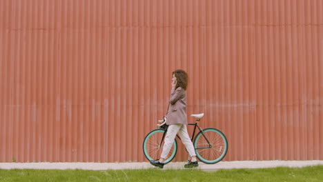 Mujer-Joven-Con-Ropa-Formal-Caminando-Con-Bicicleta-Y-Haciendo-Una-Llamada-Telefónica-Frente-A-Un-Edificio-De-Metal-Prefabricado