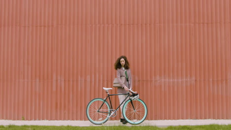 Young-Woman-In-Formal-Clothes-Walking-With-Her-Bike,-Then-Stopping-And-Looking-At-The-Camera-In-Front-Of-A-Prefab-Metal-Building