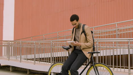 Joven-Americano-Vestido-Con-Ropa-Formal-Usando-Una-Tableta-Mientras-Se-Apoya-En-Bicicleta-Frente-A-Un-Edificio-De-Metal-Prefabricado