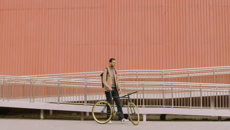 Young-American-Man-In-Formal-Clothes-Looking-At-The-Camera-While-Standing-With-His-Bike-In-Front-Of-A-Prefab-Metal-Building