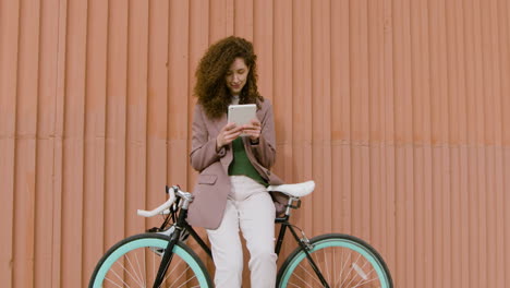 Curly-Woman-In-Formal-Clothes-Using-A-Tablet-While-Leaning-On-Bike-In-Front-Of-A-Prefab-Metal-Building
