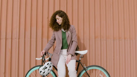 Smiling-Curly-Girl-In-Formal-Clothes-Looking-At-The-Camera-While-Leaning-On-Bike-In-Front-Of-A-Prefab-Metal-Building