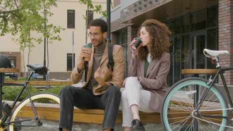 Junger-Amerikaner-Und-Frau-In-Formeller-Kleidung,-Die-Kaffee-Zum-Mitnehmen-Halten-Und-Sich-Unterhalten,-Während-Sie-Neben-Ihren-Fahrrädern-Auf-Einer-Holzbank-In-Der-Stadt-Sitzen