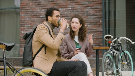 Young-American-Man-And-Woman-In-Formal-Clothes-Drinking-Coffee-And-Talking-While-Sitting-Next-To-Their-Bikes-On-A-Wooden-Bench-In-The-City