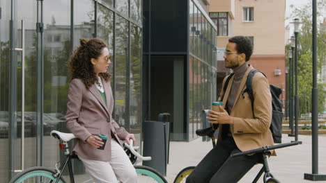 Young-American-Man-And-Woman-In-Formal-Clothes-Drinking-Coffee-And-Talking-While-Leaning-On-Their-Bikes-In-The-Street