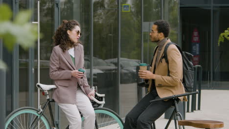Young-American-Man-And-Woman-In-Formal-Clothes-Drinking-Coffee-And-Talking-While-Leaning-On-Their-Bikes-In-The-Street-1