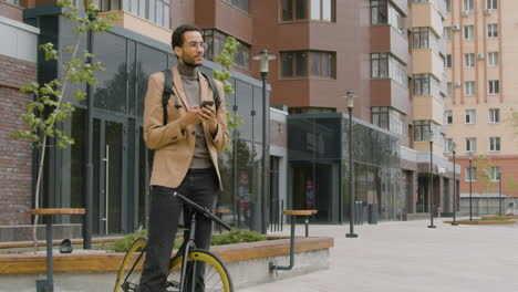Smiling-American-Man-In-Formal-Clothes-Using-Mobile-Phone-While-Standing-With-His-Bike-In-The-Street