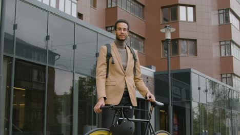 Young-American-Man-In-Formal-Clothes-And-Backpack-Standing-In-The-Street-With-His-Bike-And-Looking-At-The-Camera