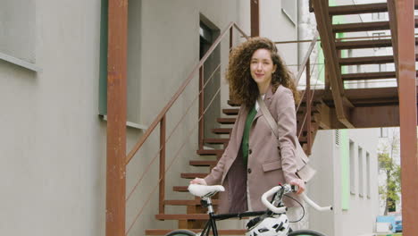 Beautiful-Curly-Woman-In-Formal-Clothes-Looking-And-Smiling-At-The-Camera-While-Standing-With-Her-Bike-Outside-In-The-City