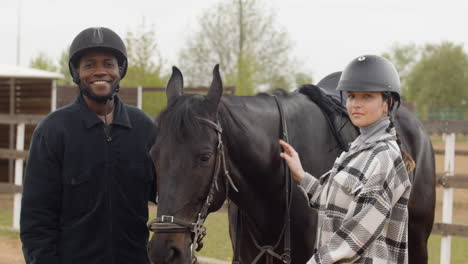 Two-Friends-Petting-Black-Horse-And-Looking-At-Camera-Outdoors