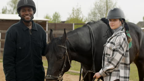 Two-Friends-Petting-Black-Horse-And-Looking-At-Camera-Outdoors-1