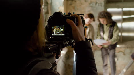 Rear-View-Of-Cameraman-Recording-His-Coworkers-Reading-A-Script-In-A-Ruined-Building