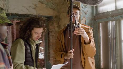Three-Production-Coworkers-Talking-While-Reading-A-Document-And-Holding-A-Microphone-During-Recording-Of-A-Movie-In-A-Ruined-Building