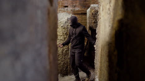 Masked-Man-Wearing-Black-Clothes-Holding-An-Ax-While-Walking-In-A-Ruined-Building