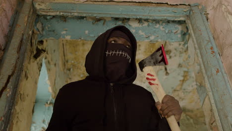 Bottom-View-Of-Man-Wearing-Black-Balaclava-And-Holding-An-Ax-In-A-Ruined-Building